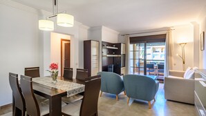 Uma sala de estar moderna com área de jantar, apresentando uma mesa grande, cadeiras, um sofá e uma cozinha com armários e uma geladeira.