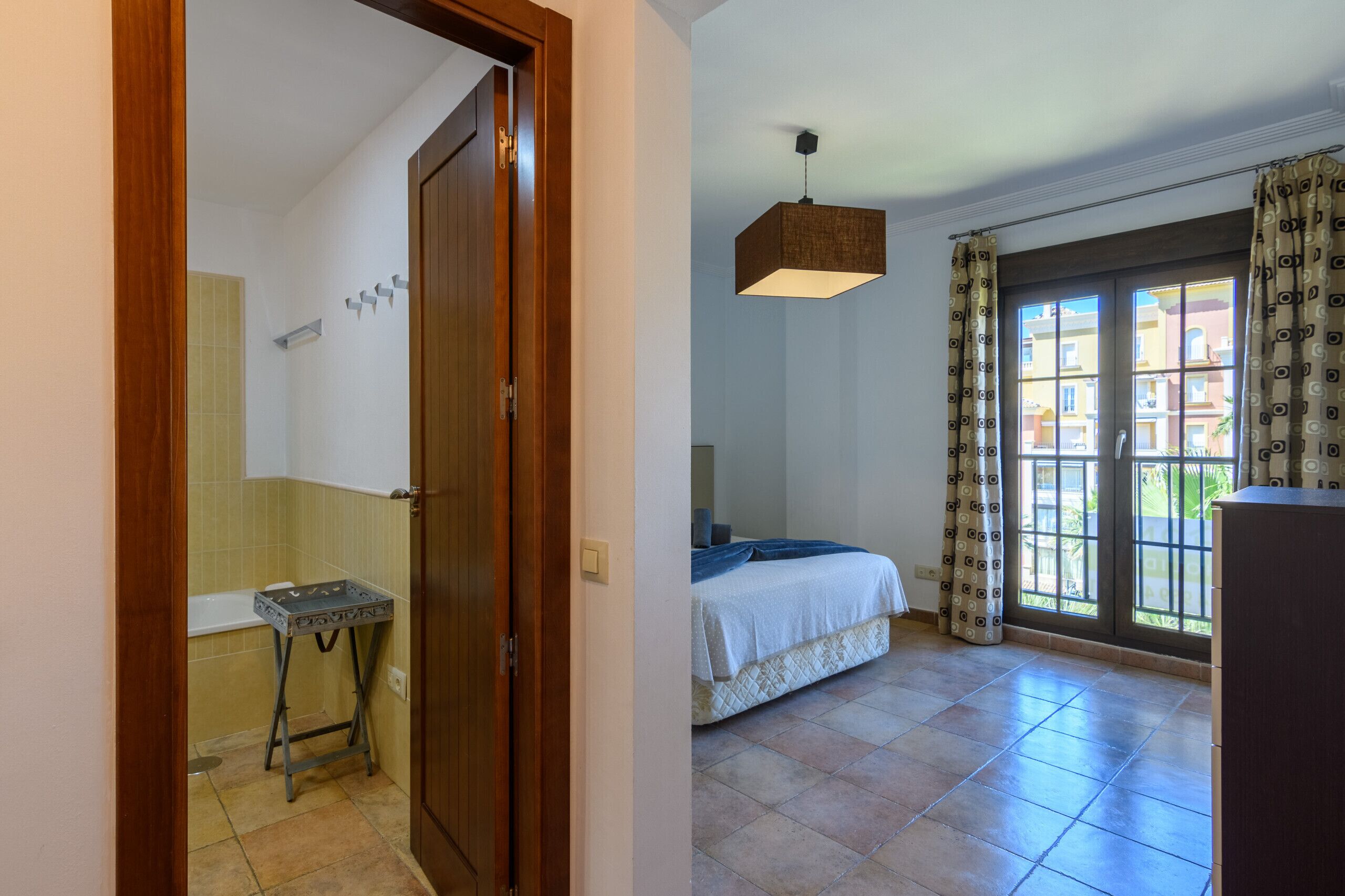 A bedroom with a bed, a window with curtains, and a bathroom with a bathtub and a towel rack.