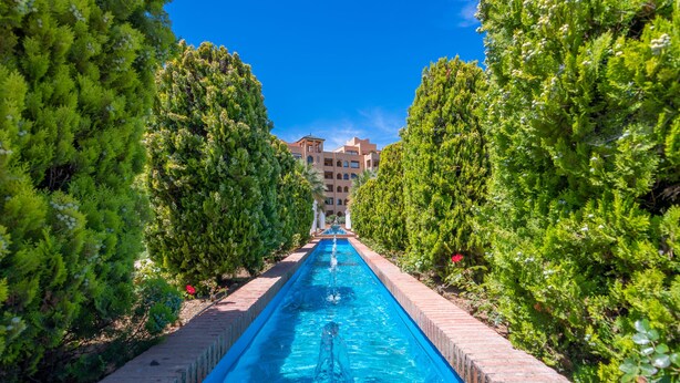 Uma piscina comprida e estreita ladeada por árvores verdes exuberantes, conduzindo a um edifício com vários andares.