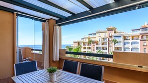 Varanda com mesa e cadeiras, um vaso de planta e vista para um edifício com várias andares e varandas.