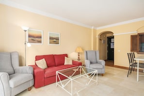 Un salon avec un canapé rouge, deux fauteuils, une table basse en verre et une lampe.