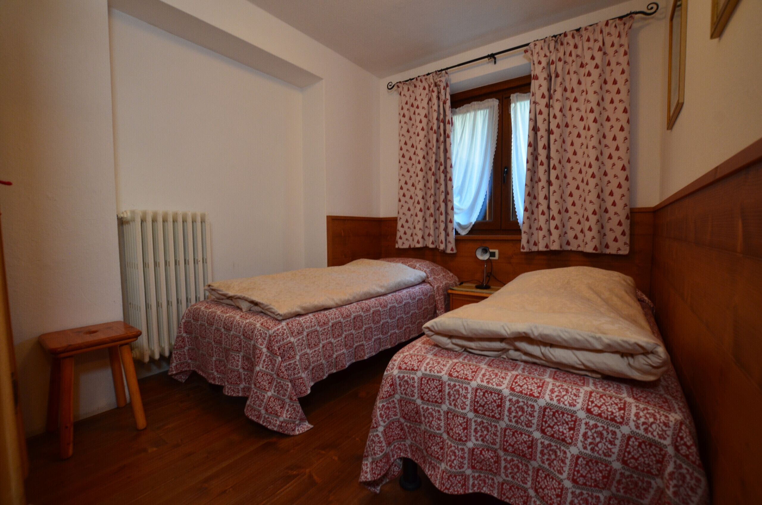 Une petite chambre avec deux lits simples, une table de chevet en bois, un radiateur et une fenêtre avec des rideaux.