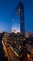 Un edificio de gran altura con fachada de vidrio moderna, rodeado de otros edificios y una calle concurrida al anochecer.