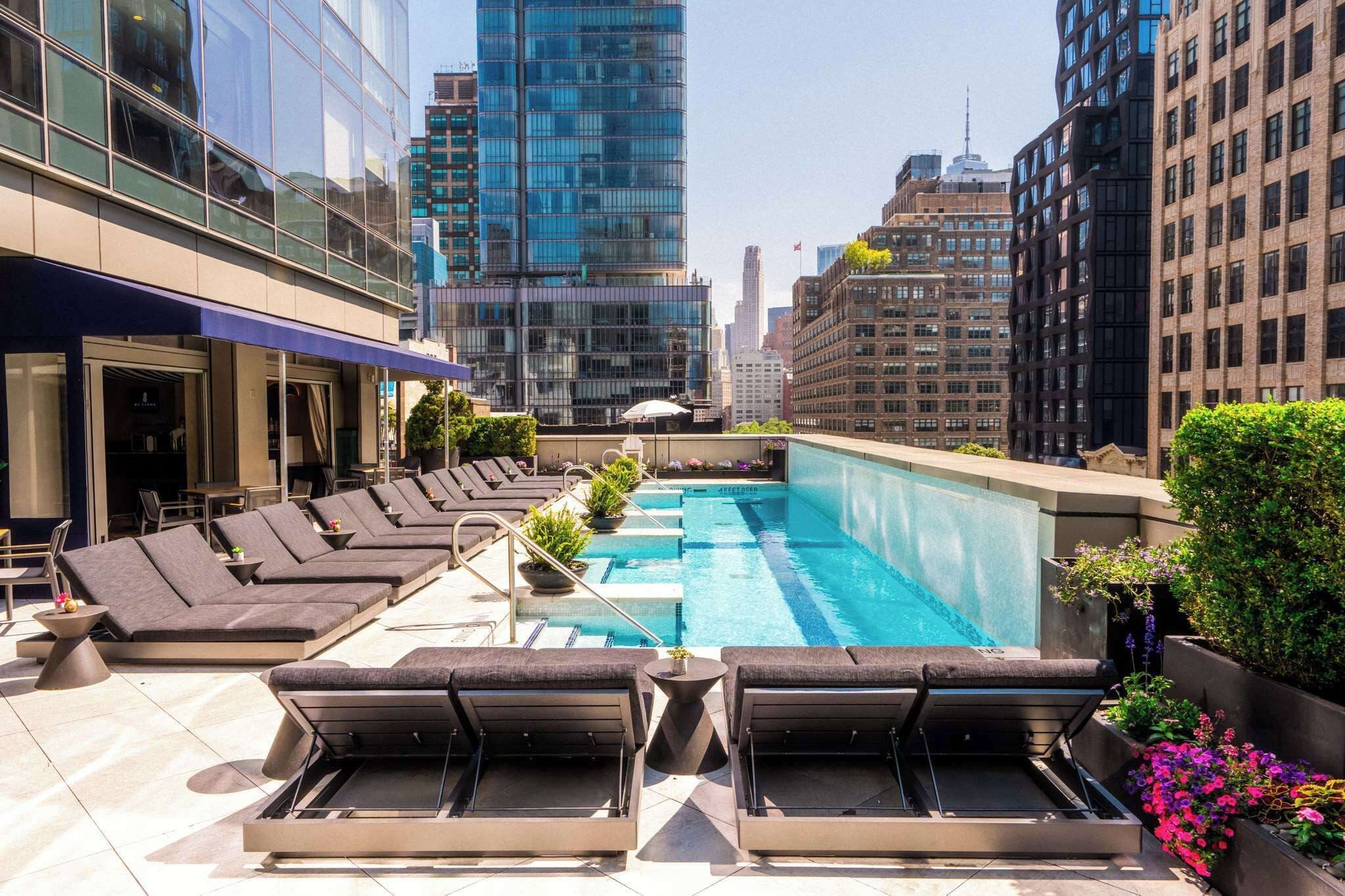 Área de piscina no terraço, com espreguiçadeiras, piscina e edifícios ao redor.
