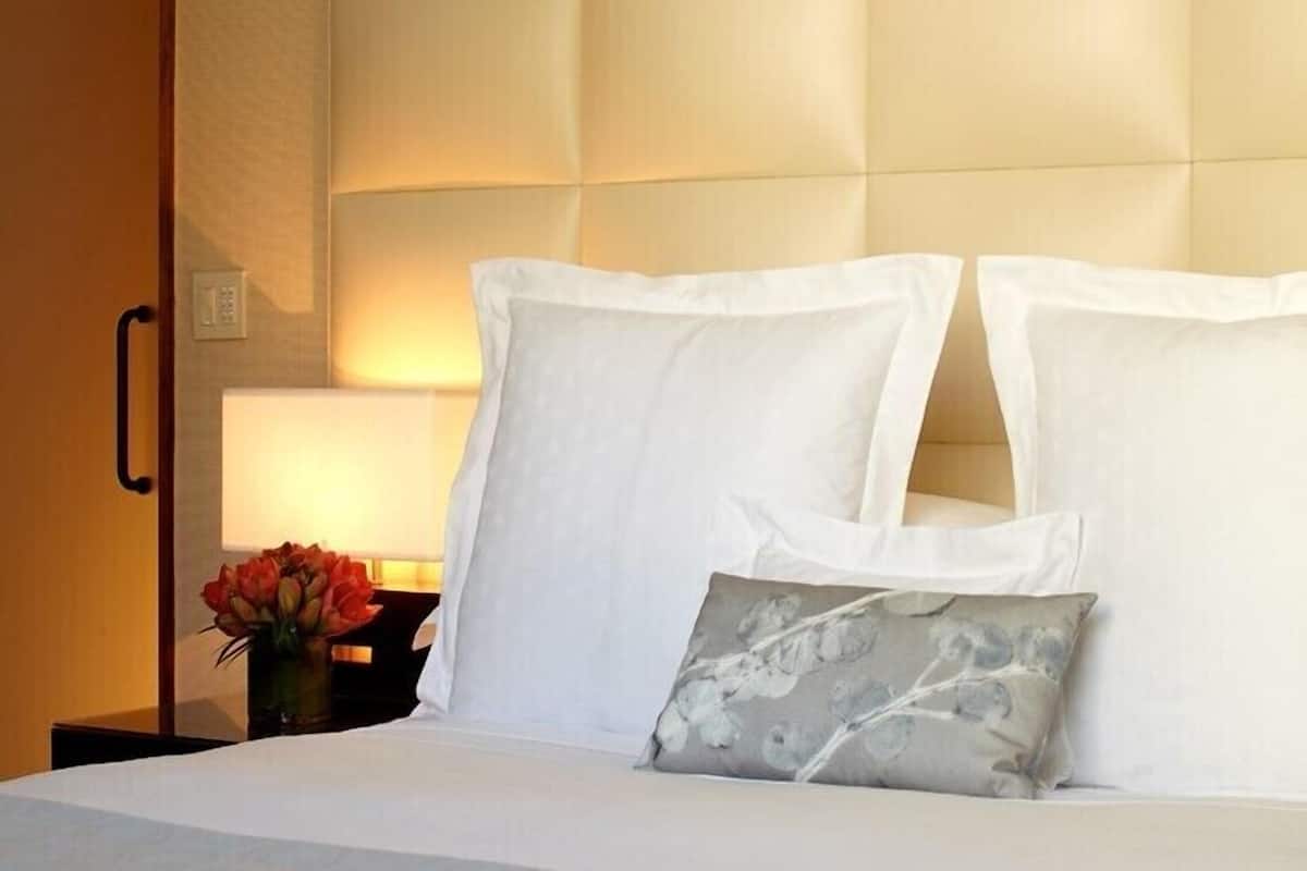 A hotel room with a bed, white pillows, a grey floral pillow, and a nightstand with a vase of flowers.