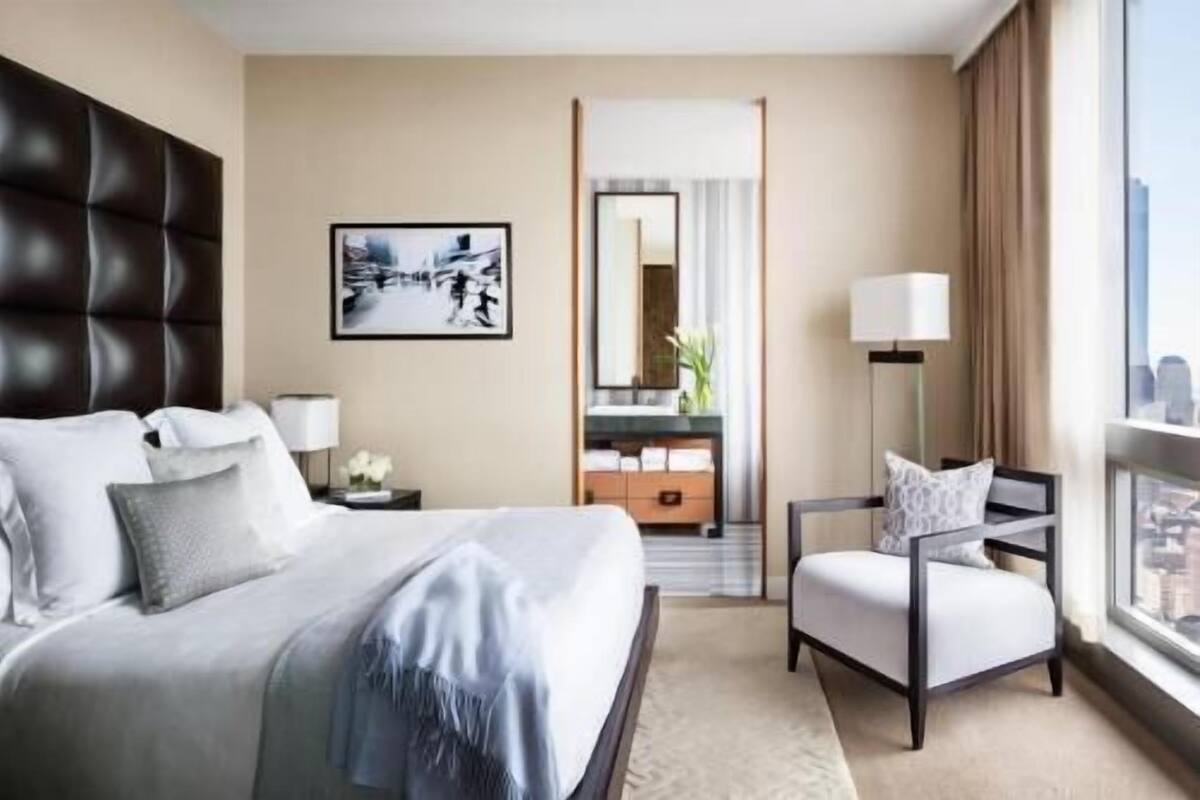 A hotel room with a large bed, a chair, a desk, a mirror, and a window with a city view.