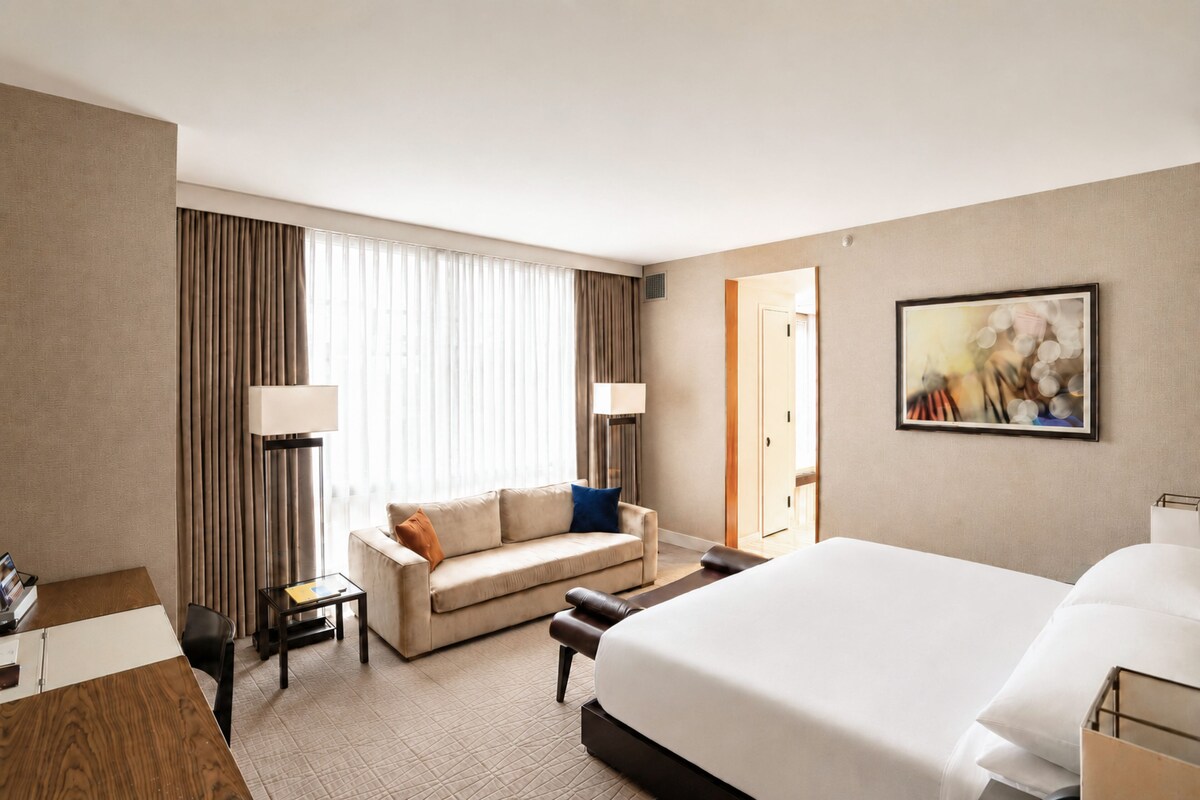 Quarto de hotel com uma cama grande, um sofá, uma escrivaninha e uma cadeira. Há uma janela com cortinas, uma porta e um quadro na parede.