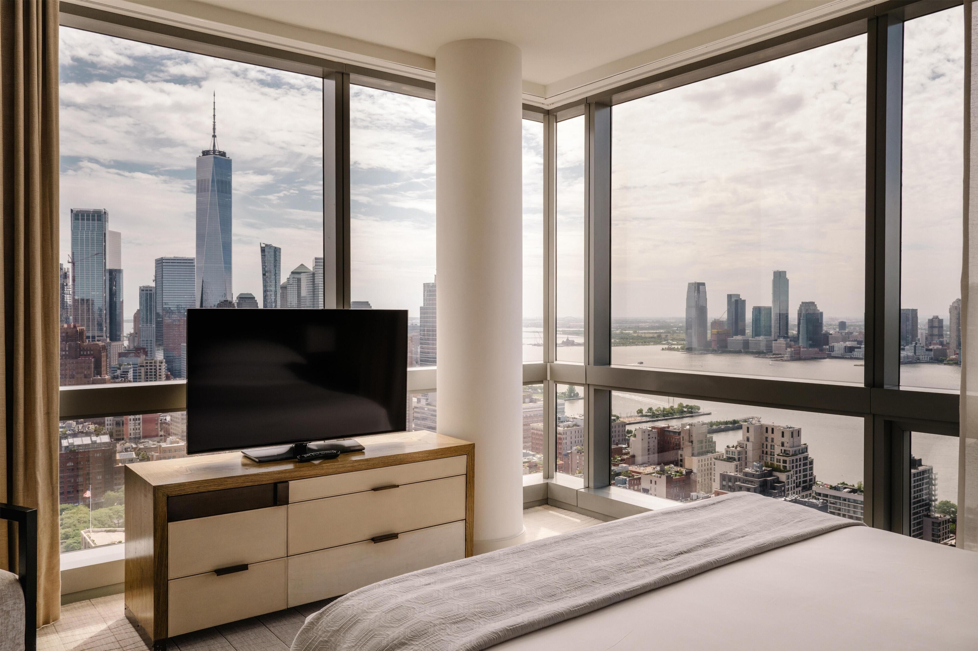 Une chambre avec une grande fenêtre, un téléviseur à écran plat, une commode en bois et un lit.