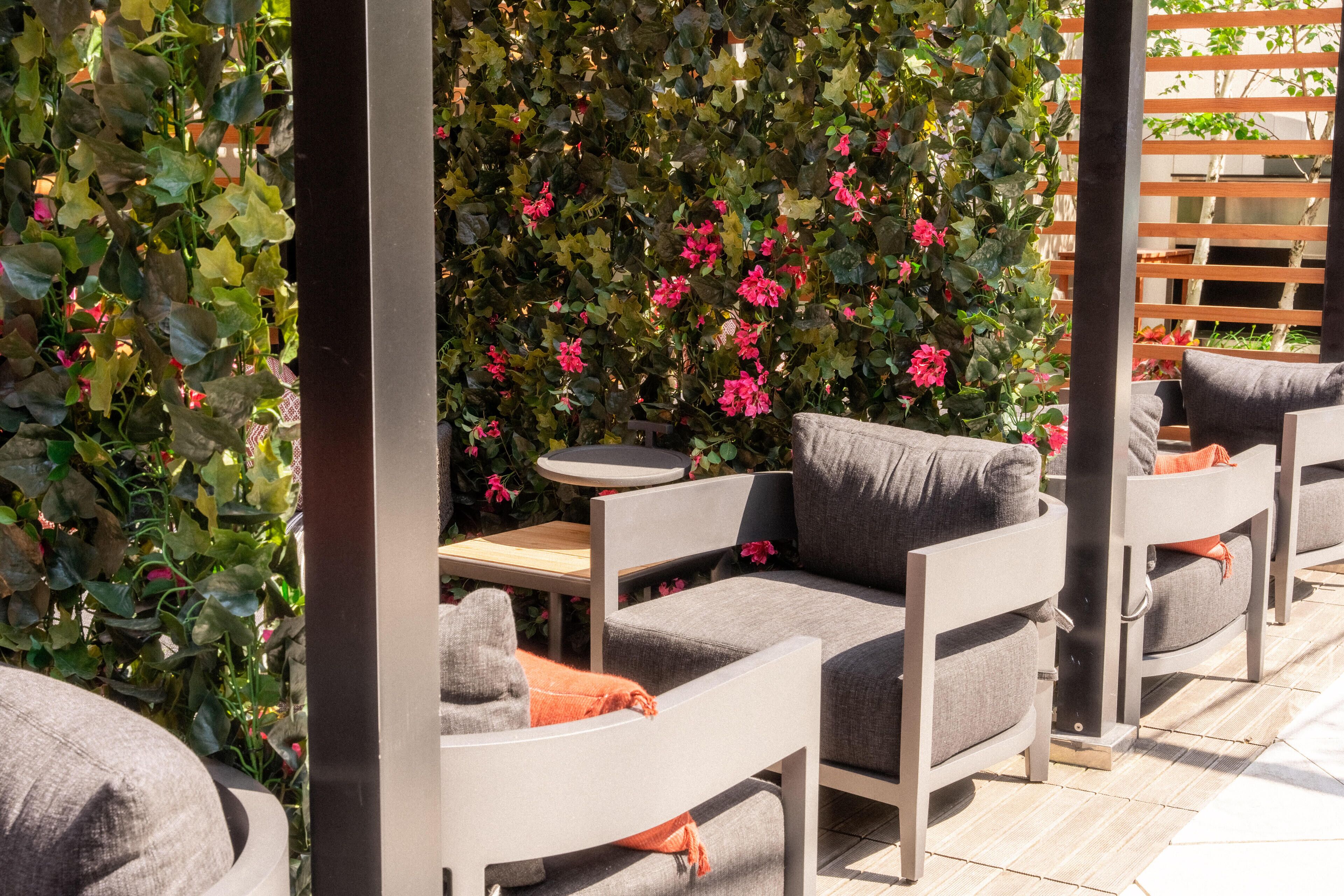 An outdoor seating area with grey cushioned chairs and a small round table, surrounded by lush green foliage and vibrant pink flowers.