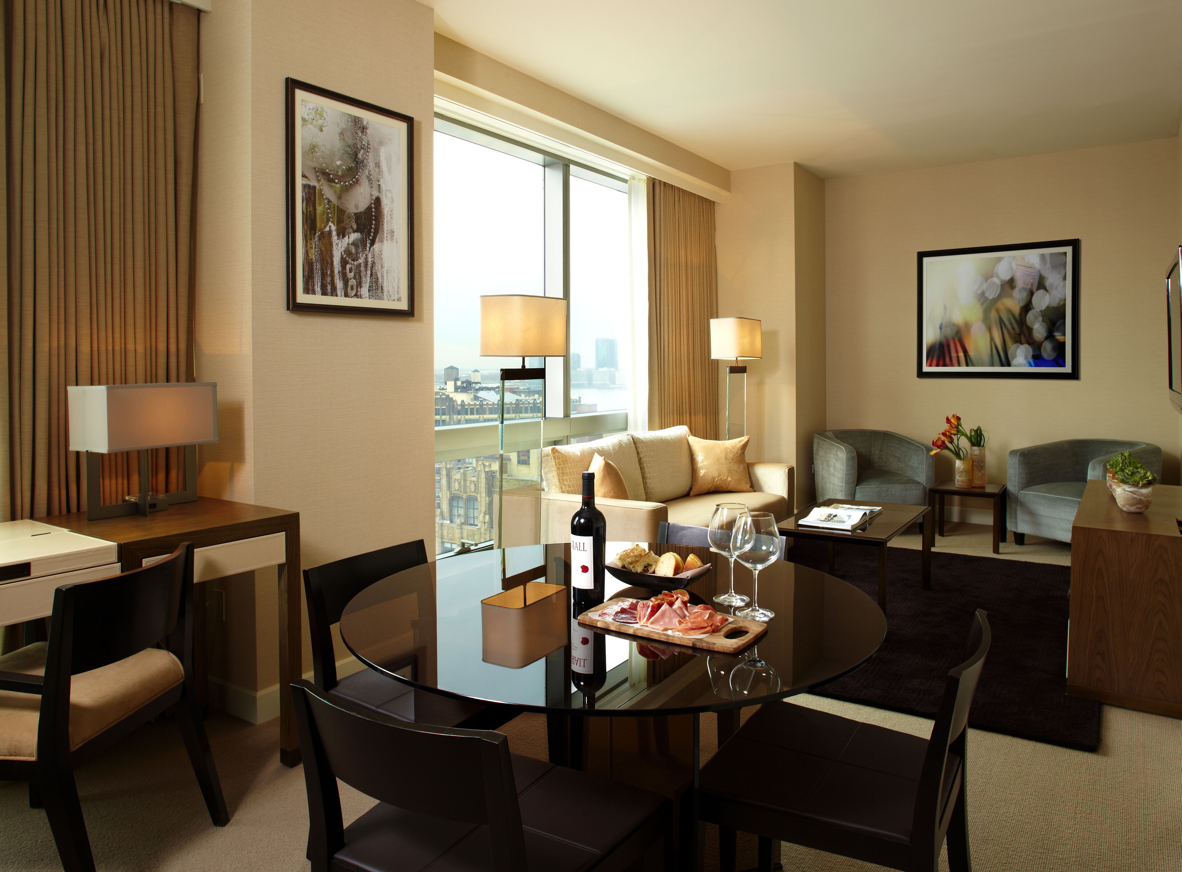 Hotelkamer met een eethoek, een bank, een stoel, een lamp en een raam met uitzicht op de stad.