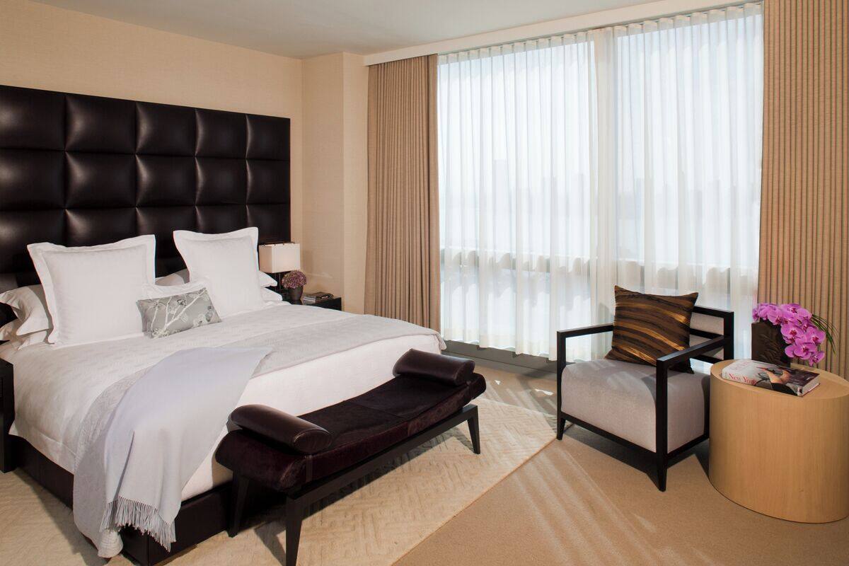 A modern hotel room with a large bed, a dark upholstered bench, a side table with a magazine, and a window with sheer curtains.