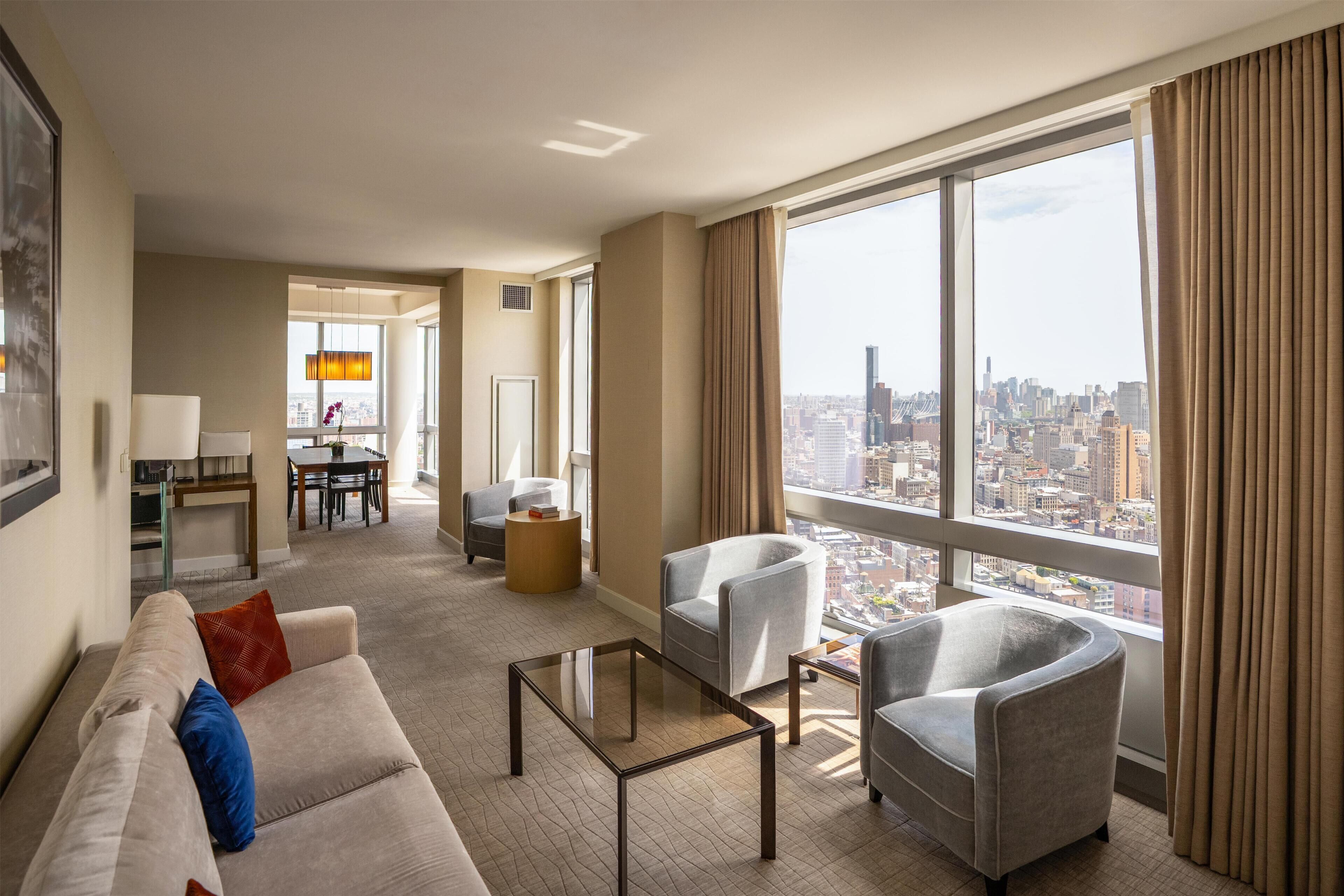 A hotel room with a large window, a sofa, two armchairs, a coffee table, and a dining area with a table and chairs.