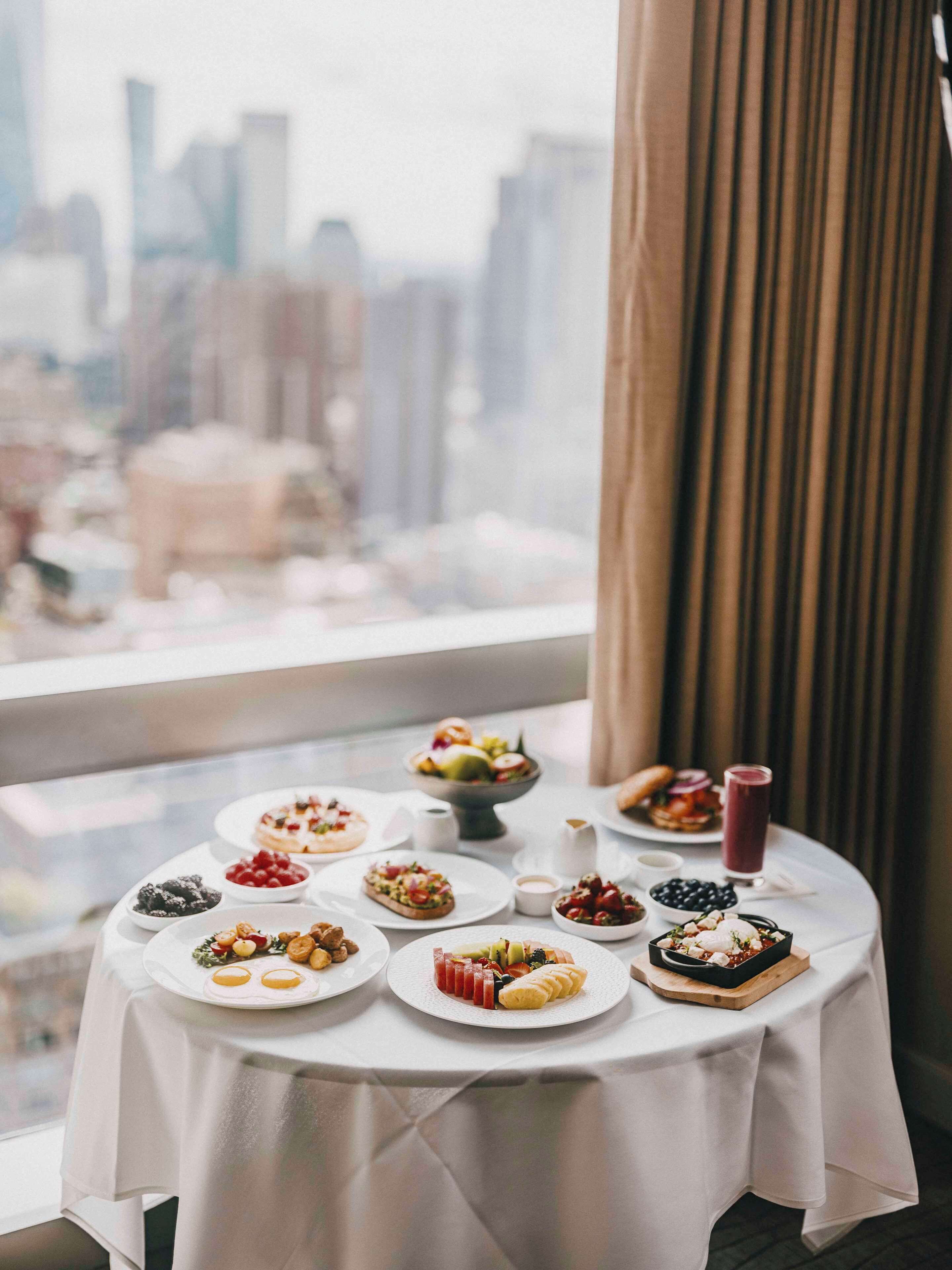 A round table with various dishes and a glass of red beverage, set against a window with a cityscape view.