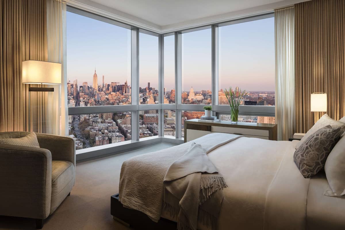 A hotel room with a large window offering a city view, a bed, a desk, and a chair.