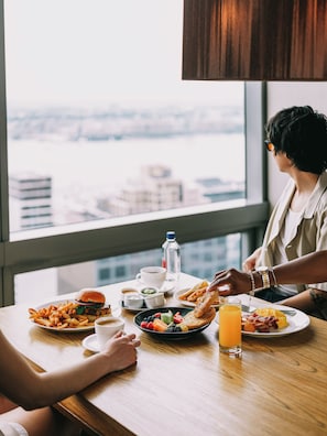 A dining table with various breakfast items, a city view through a window, and a person reaching for a plate.