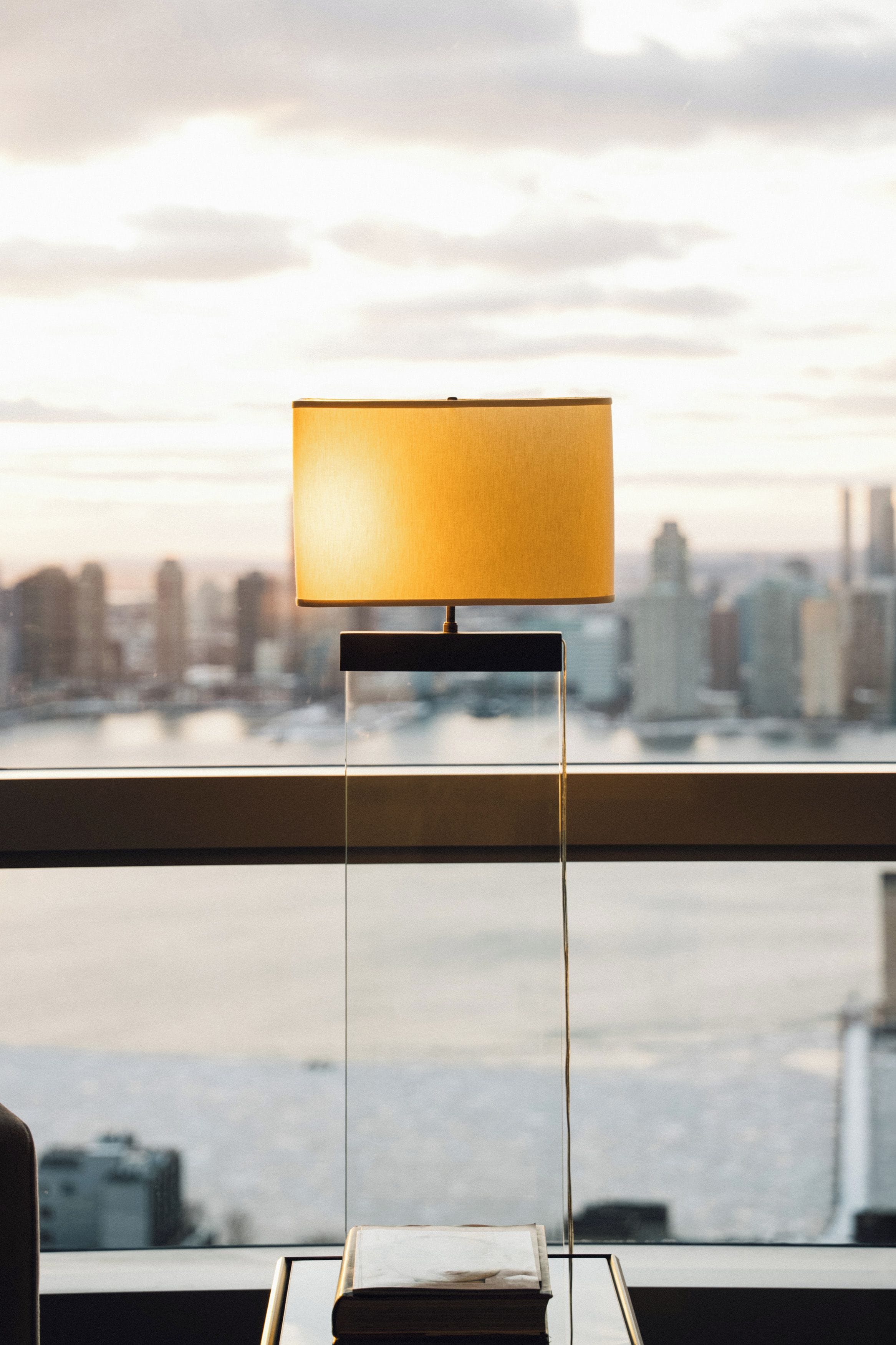 A room with a view of a city skyline through a window, a table with a lamp, and a book on the table.