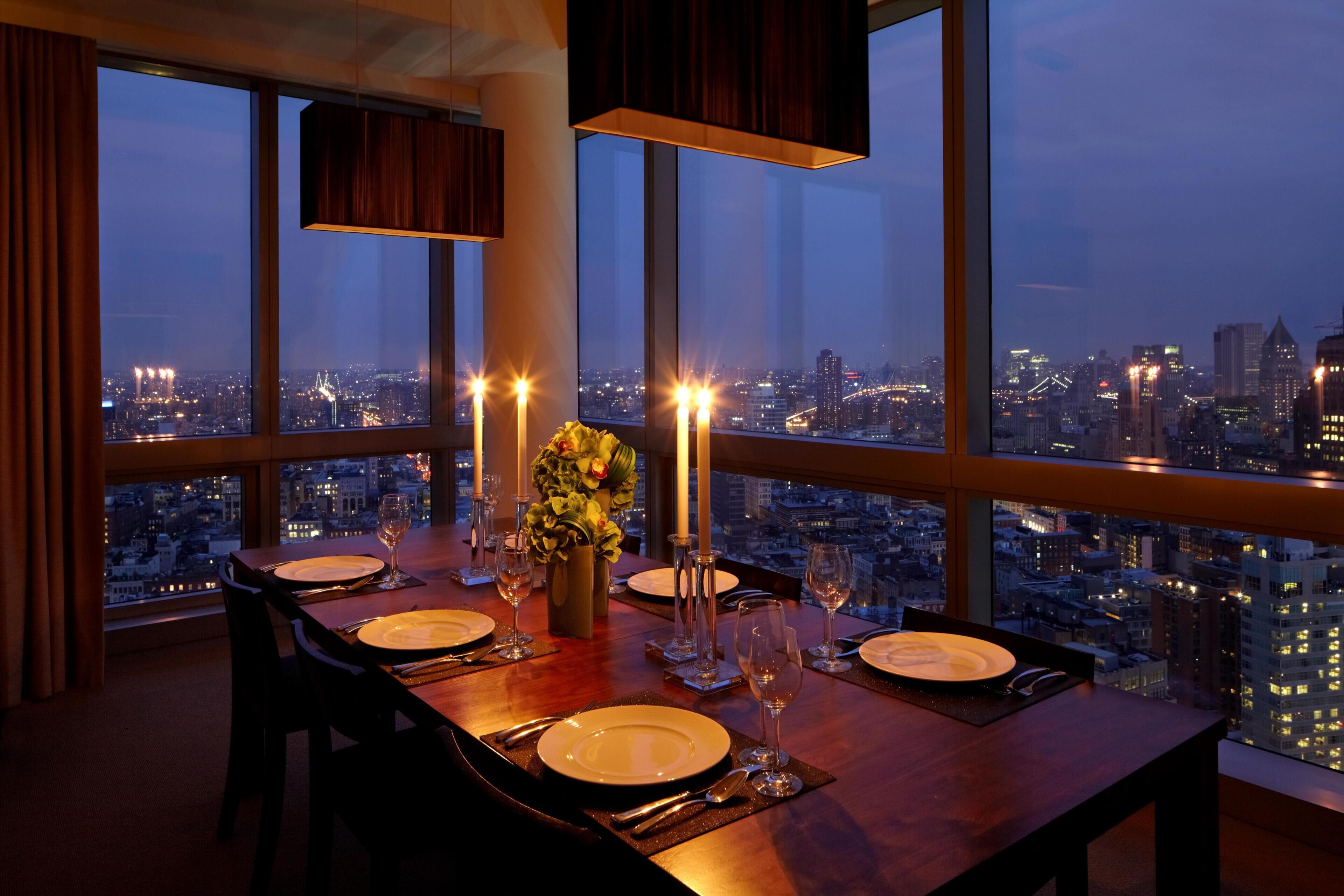 A dining table set for four with candles, a vase of flowers, and a city view.