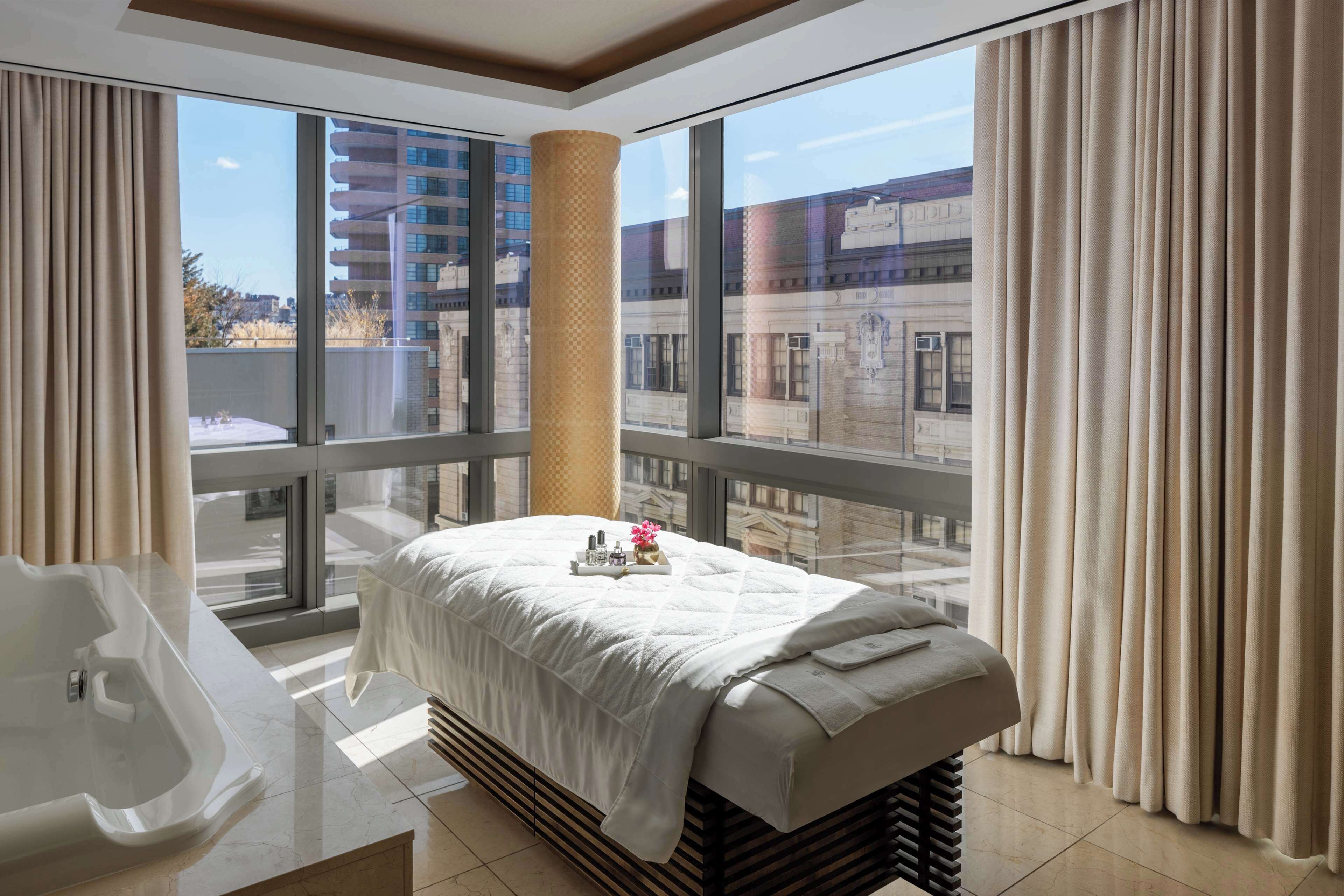 Habitación de hotel con una cama grande, una camilla de masaje, una bañera y vistas a la ciudad a través de amplios ventanales.