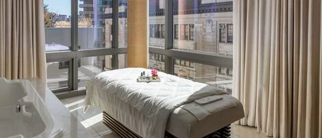 Habitación de hotel con una cama grande, una camilla de masaje, una bañera y vistas a la ciudad a través de amplios ventanales.