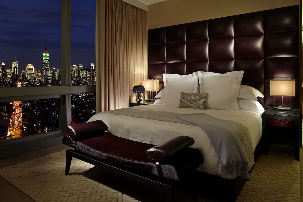 A hotel room with a large bed, two bedside tables with lamps, a view of a city skyline, and a cityscape through the window.