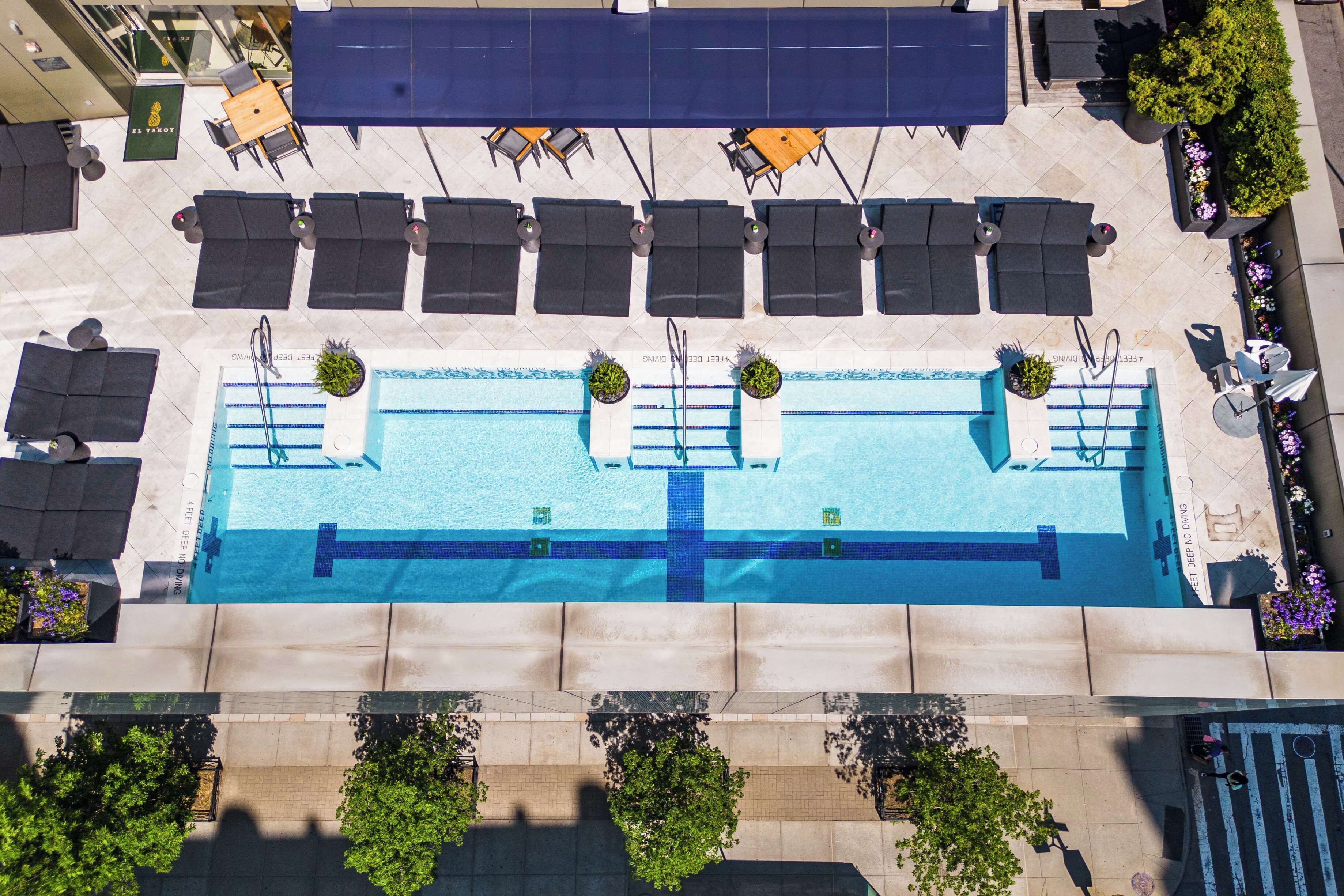 An outdoor swimming pool with lounge chairs and tables, surrounded by potted plants and a tiled walkway.