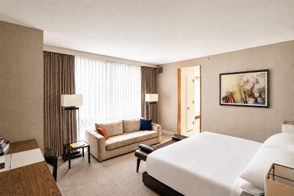 Quarto de hotel com uma cama grande, um sofá, uma escrivaninha e uma cadeira. Há uma janela com cortinas, uma porta e um quadro na parede.