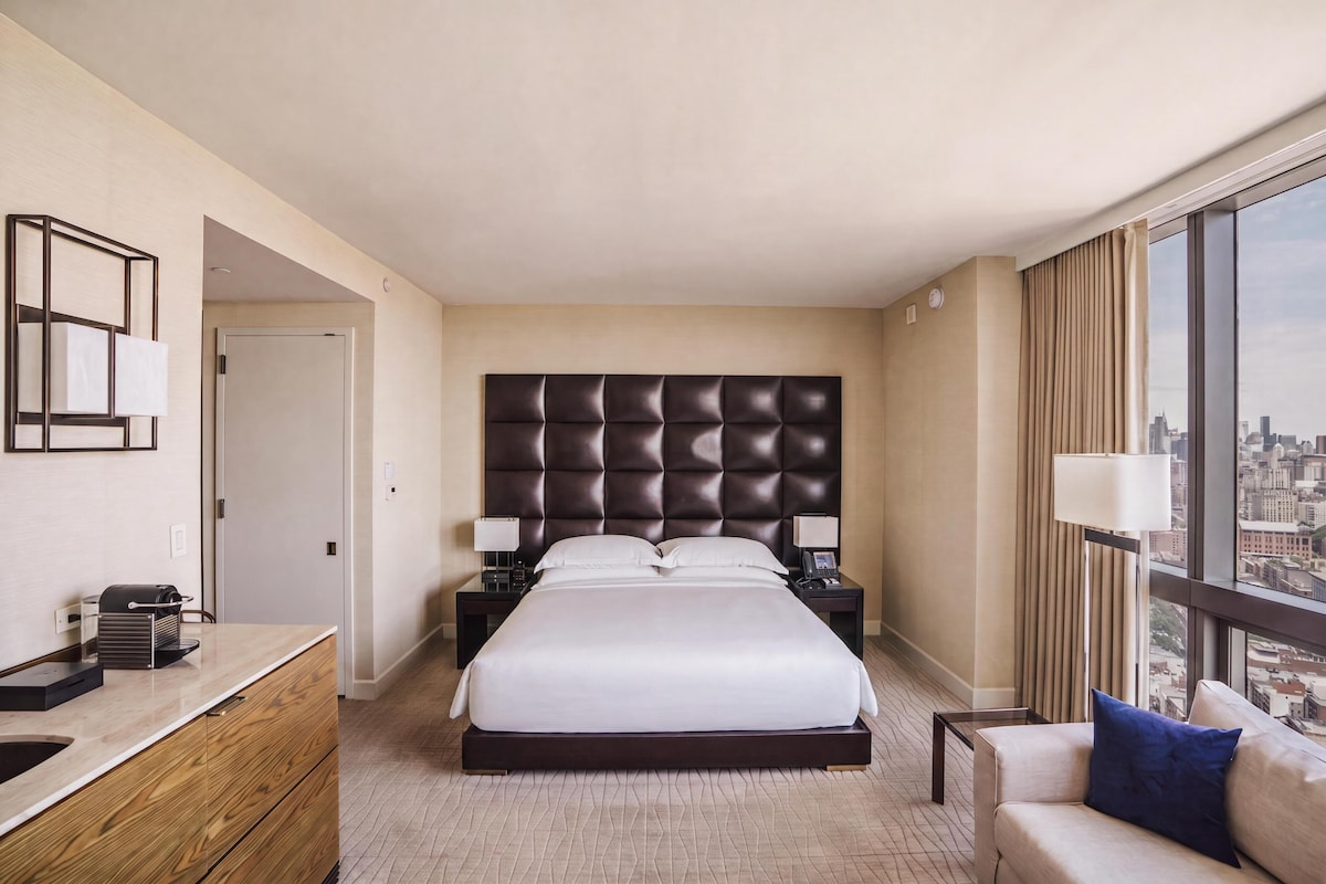 A hotel room with a large bed, a desk with a lamp, a chair, and a view of the city through the window.
