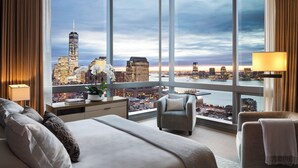 Une chambre d’hôtel avec un grand lit, deux fauteuils, un bureau avec une lampe et une vue sur le panorama de la ville.