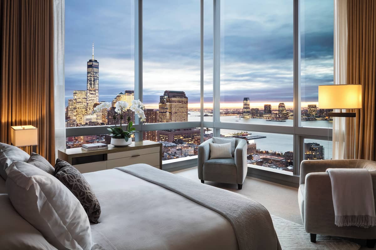 A hotel room with a large bed, two armchairs, a desk with a lamp, and a view of the city skyline.