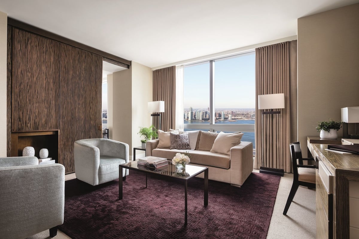 A modern hotel room with a city view, a sofa, armchairs, a coffee table, and a dining area.