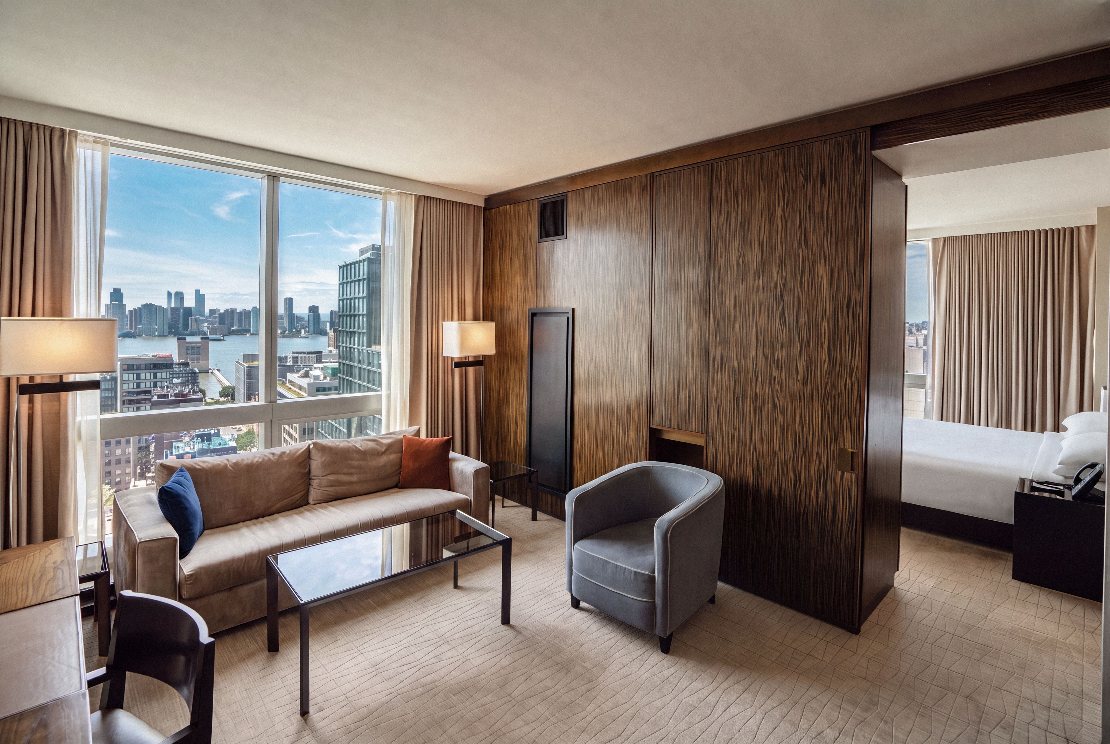 A modern hotel room with a large window, a sofa, a chair, a small table, and a bed.