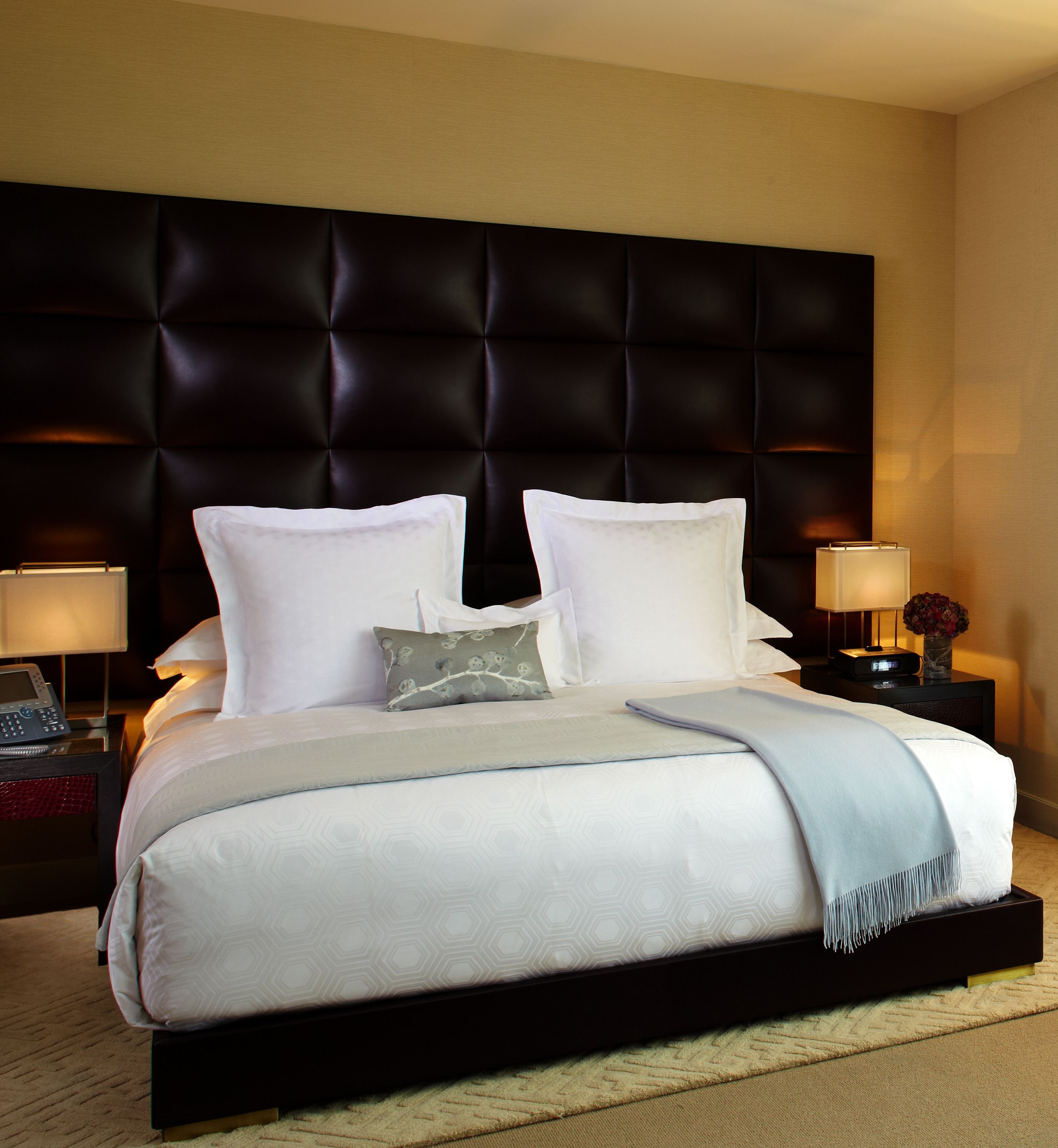 A hotel room with a large bed, a dark headboard, two bedside lamps, and a nightstand with a clock and a vase of flowers.