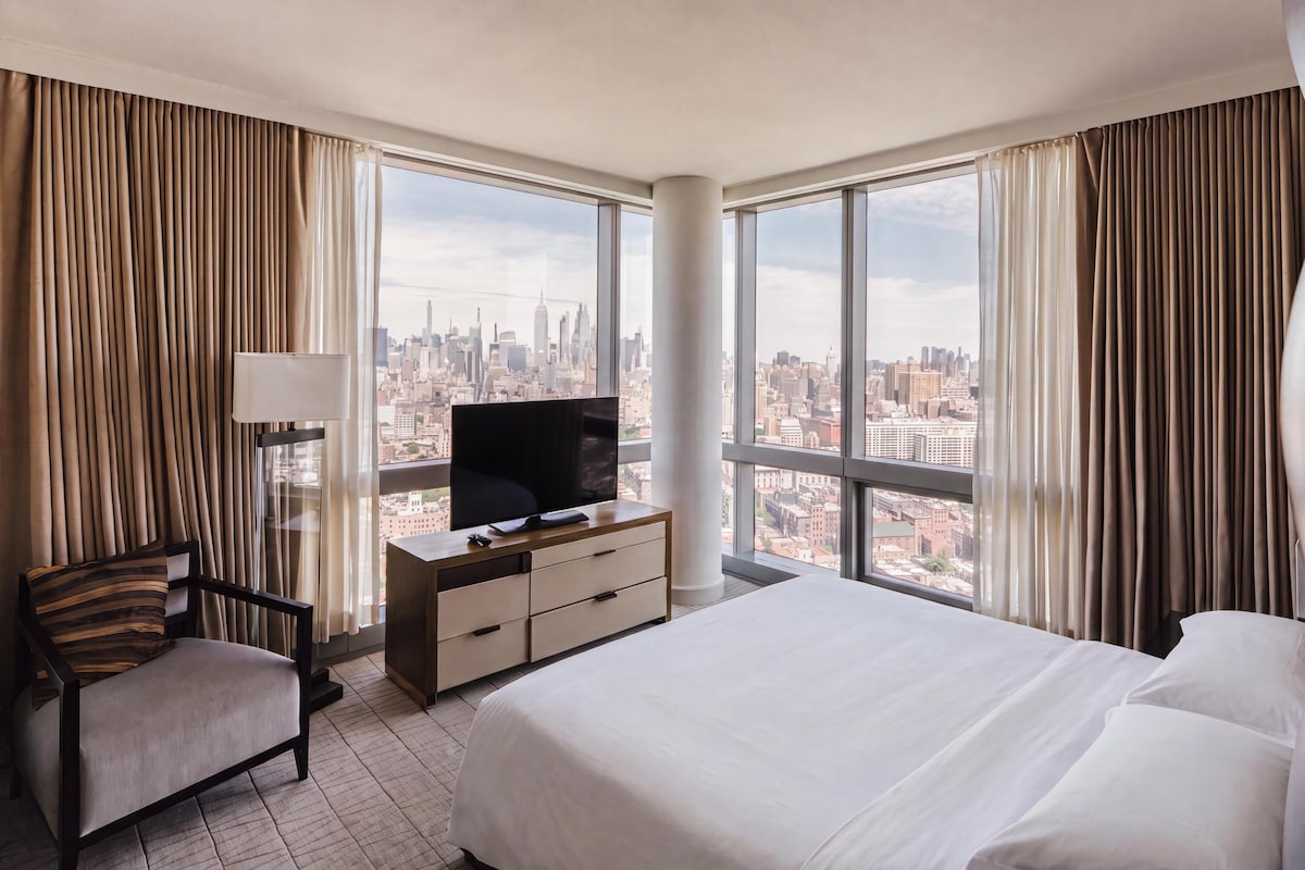 A hotel room with a large bed, a TV on a wooden dresser, a chair, and a view of a cityscape through large windows.