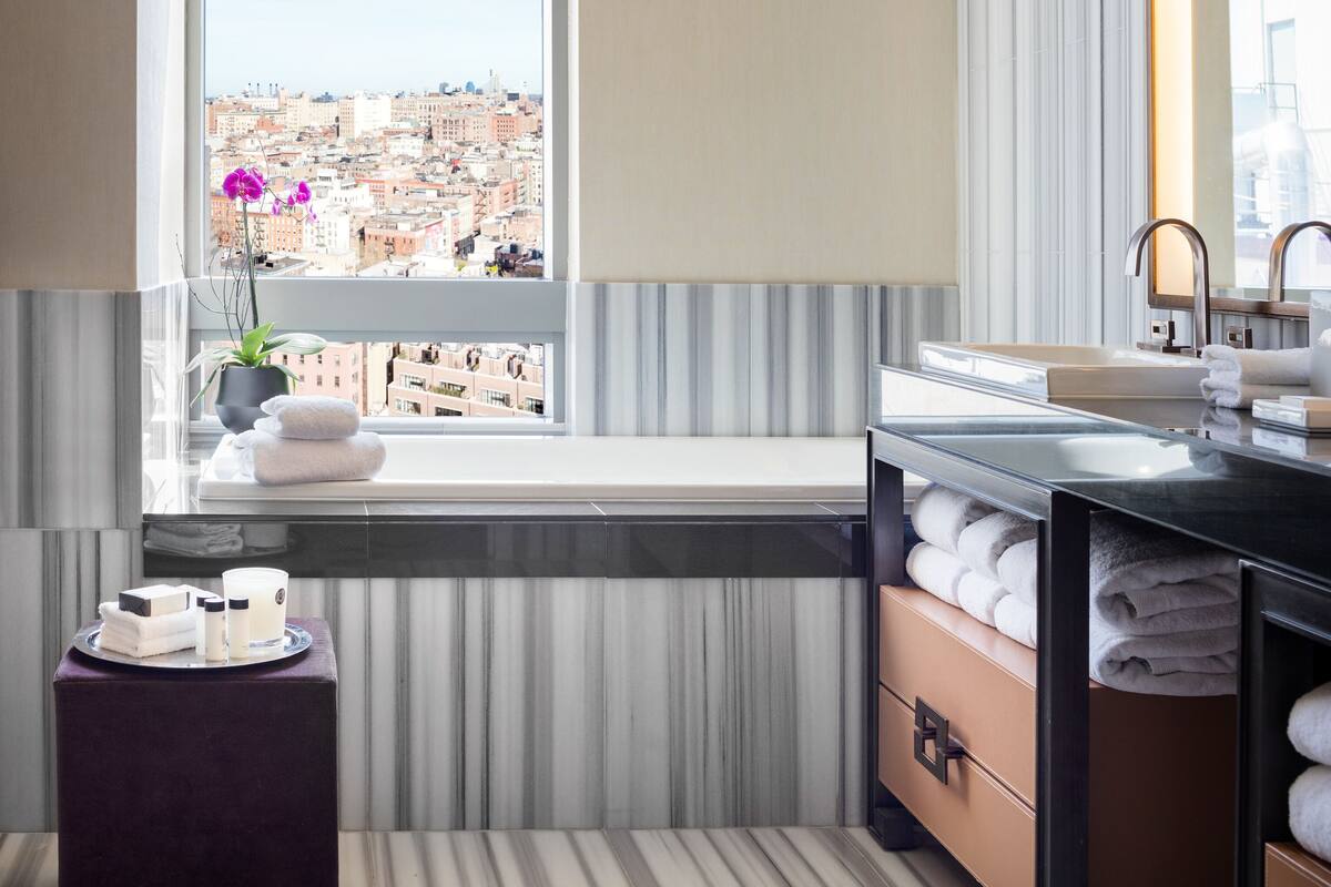A modern bathroom with a large window, a view of a cityscape, a vanity with a mirror, and neatly folded towels.