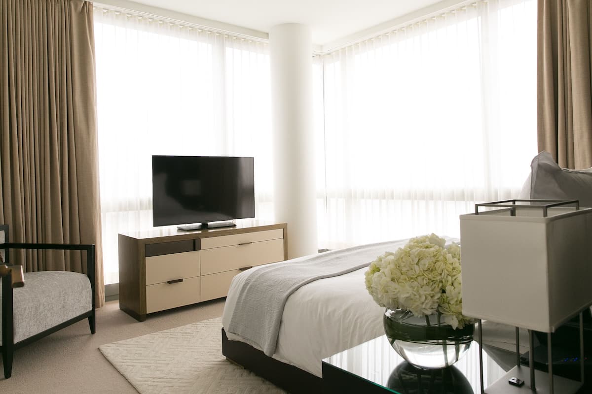 A modern hotel room with a bed, a TV, a bedside table with a vase of flowers, and a lamp.
