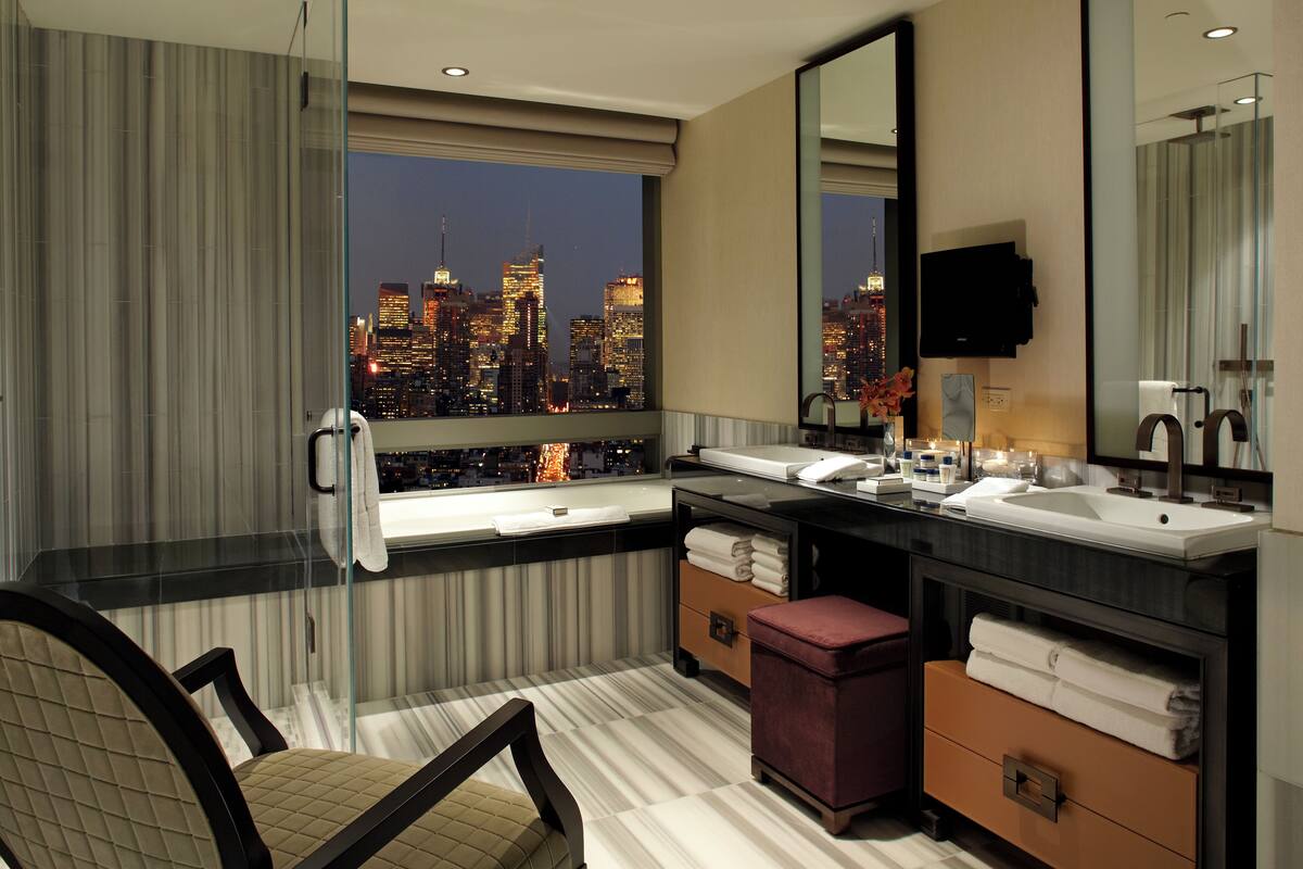 A modern bathroom with a dual sink vanity, a large mirror, and a view of a city skyline at night.