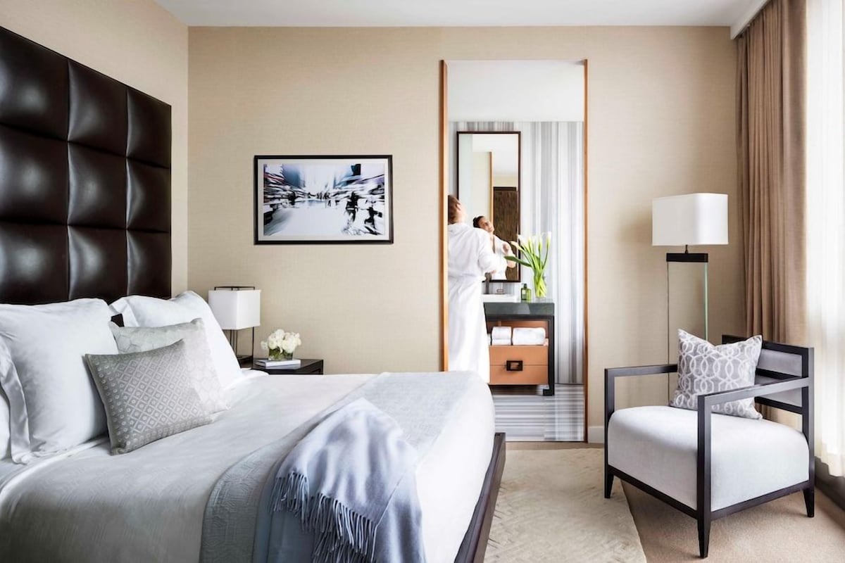 Quarto de hotel com cama, poltrona, mesa de cabeceira, abajur, espelho e um quadro pendurado na parede.