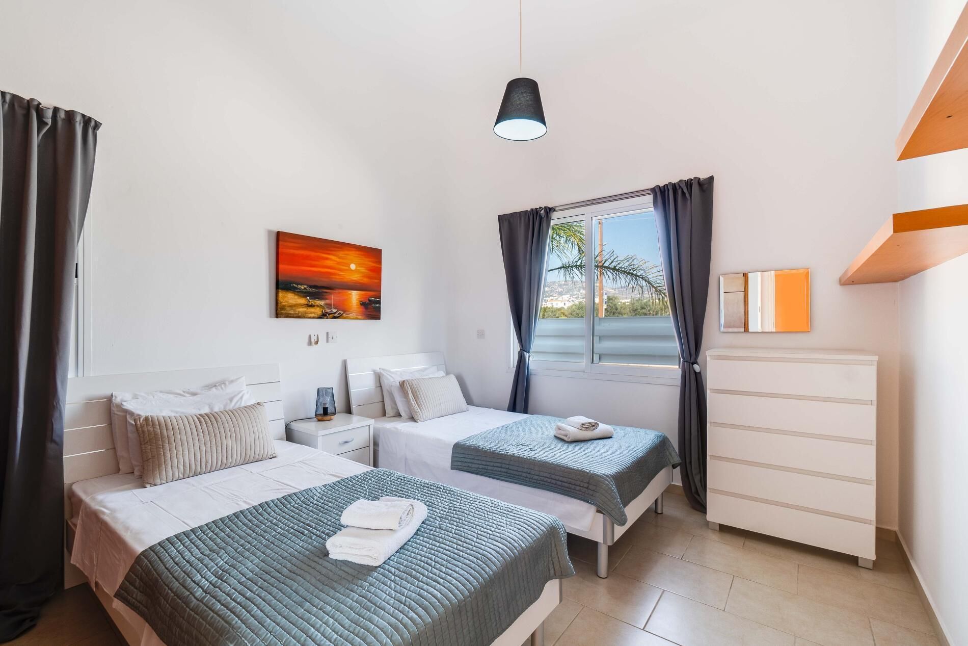 A hotel room with two beds, a nightstand, a lamp, a painting, a window with curtains, and a dresser.