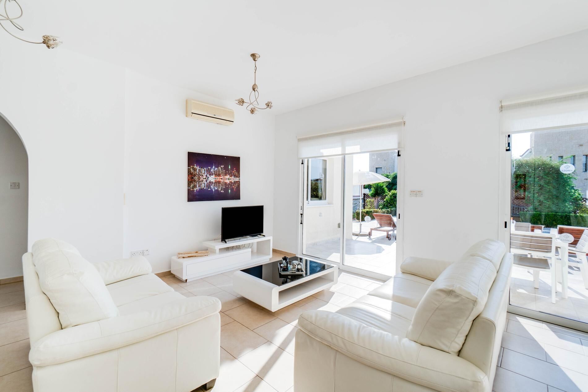 Ein modernes Wohnzimmer mit weißen Sofas, einem Flachbildfernseher und einem Couchtisch. An der Wand hängt ein Stadtbild und eine Glastür führt in einen Außenbereich.