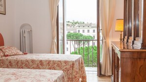 A hotel room with two beds, a wooden dresser, a lamp, and a view of buildings and greenery through the window. - Ancient Rome View Apartment (Rome)