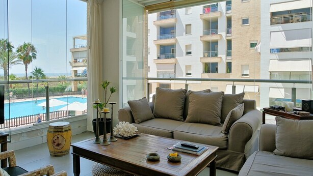 A modern living room with a large window, a sofa, a coffee table, and a view of a swimming pool and apartment buildings. - Espigon Central - Punta del Moral Beach PEHC14P79 (Punta del Moral)