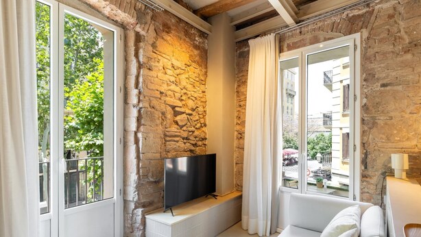 A living room with a stone wall, a flat-screen TV, a white sofa, and a view of a courtyard with trees and buildings. - Studio with terrace in Born (Barcelona)