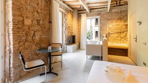 A room with stone walls, a bed with white linens, a small table with a chair, a TV, and a bathroom with a stone wall. - Studio with terrace in Born (Barcelona)
