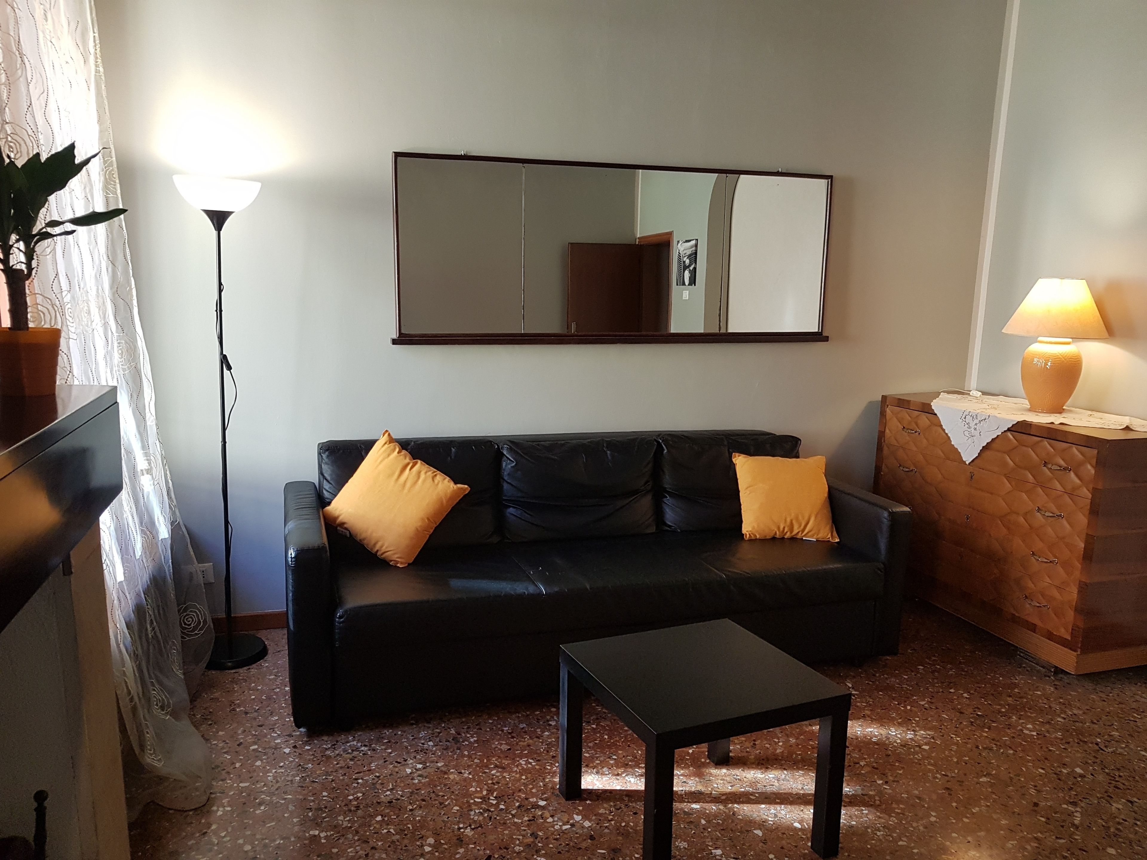 A living room with a black leather sofa, two yellow cushions, a small black table, a wooden cabinet with a lamp, and a large mirror on the wall.