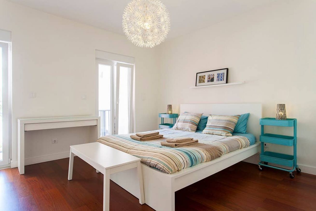 Un dormitorio con cama, mesitas de noche, un escritorio, una lámpara colgante y una ventana.
