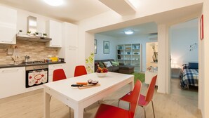 A modern kitchen with white cabinets, a dining table with red chairs, and a living area with a sofa and shelves. - Large and completely refurbished apartment with a beautiful patio. (Firenze)