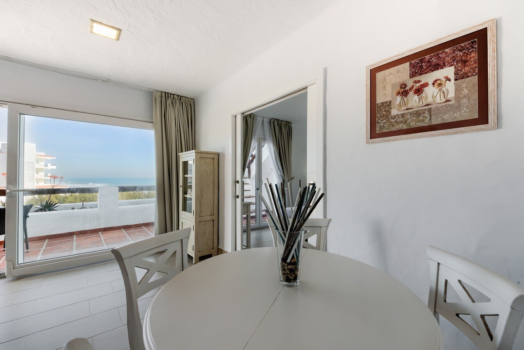 A room with a balcony, a dining table, chairs, a cabinet, and a framed picture on the wall.