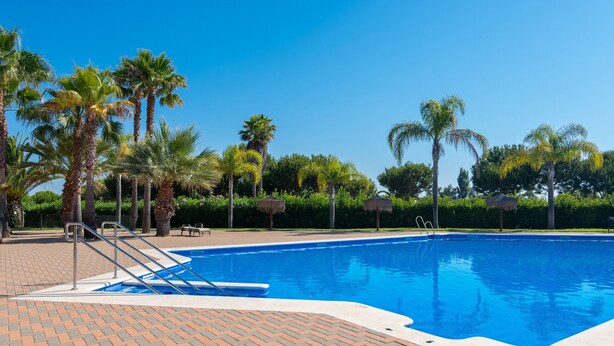A swimming pool with a brick deck, palm trees, and a clear blue sky. - Prado Golf - Isla Canela Golf GPHG45P86 (Isla Canela)