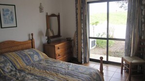 Een slaapkamer met een bed, een dressoir, een spiegel en een stoel. Er is een raam met gordijnen en uitzicht op groen.