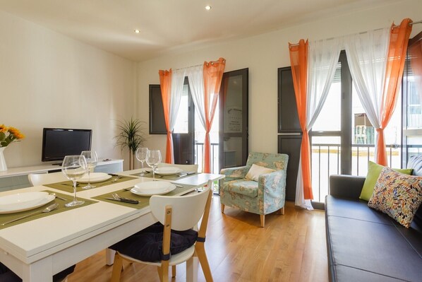 A modern living room with a dining area, featuring a sofa, a chair, a table set for two, and a television.